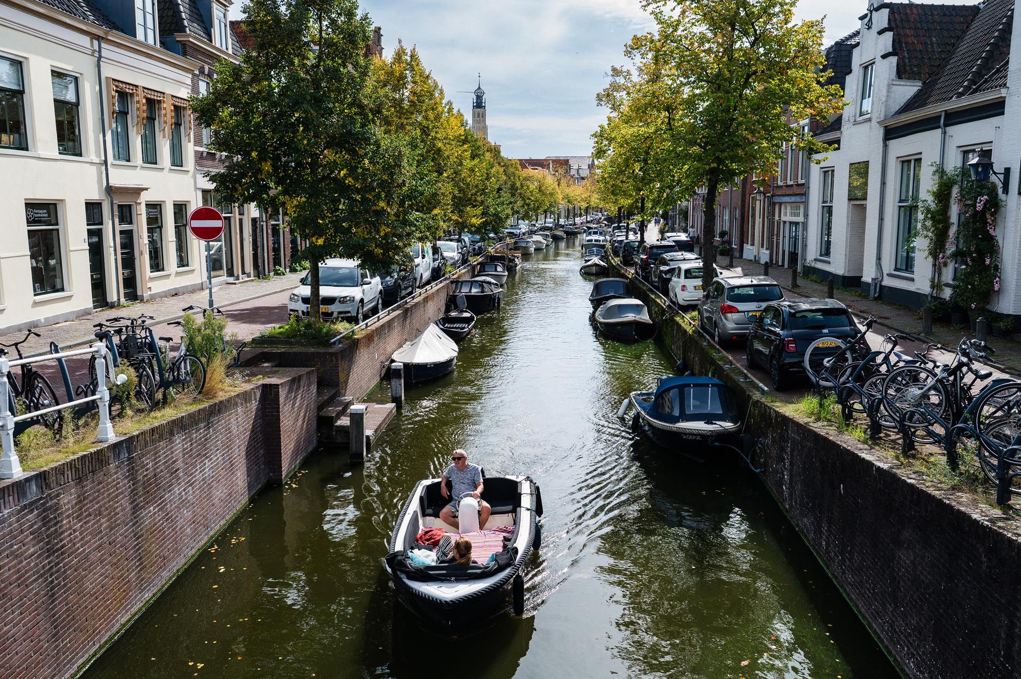 A scenic Dutch canal with a small white boat, lined with trees, bicycles, and traditional buildings on a cloudy day.