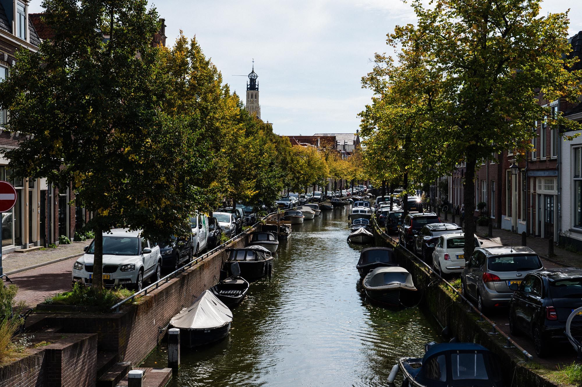 Tree-lined canal in a Dutch town with moored boats and parked cars on both sides, church tower visible in background.