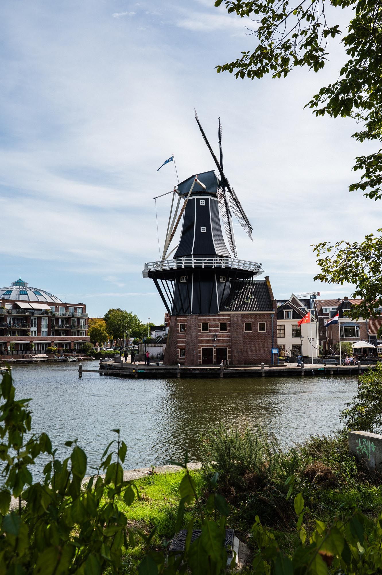 Historic Dutch windmill on a canal waterfront in Haarlem, Netherlands, under a partly cloudy sky.