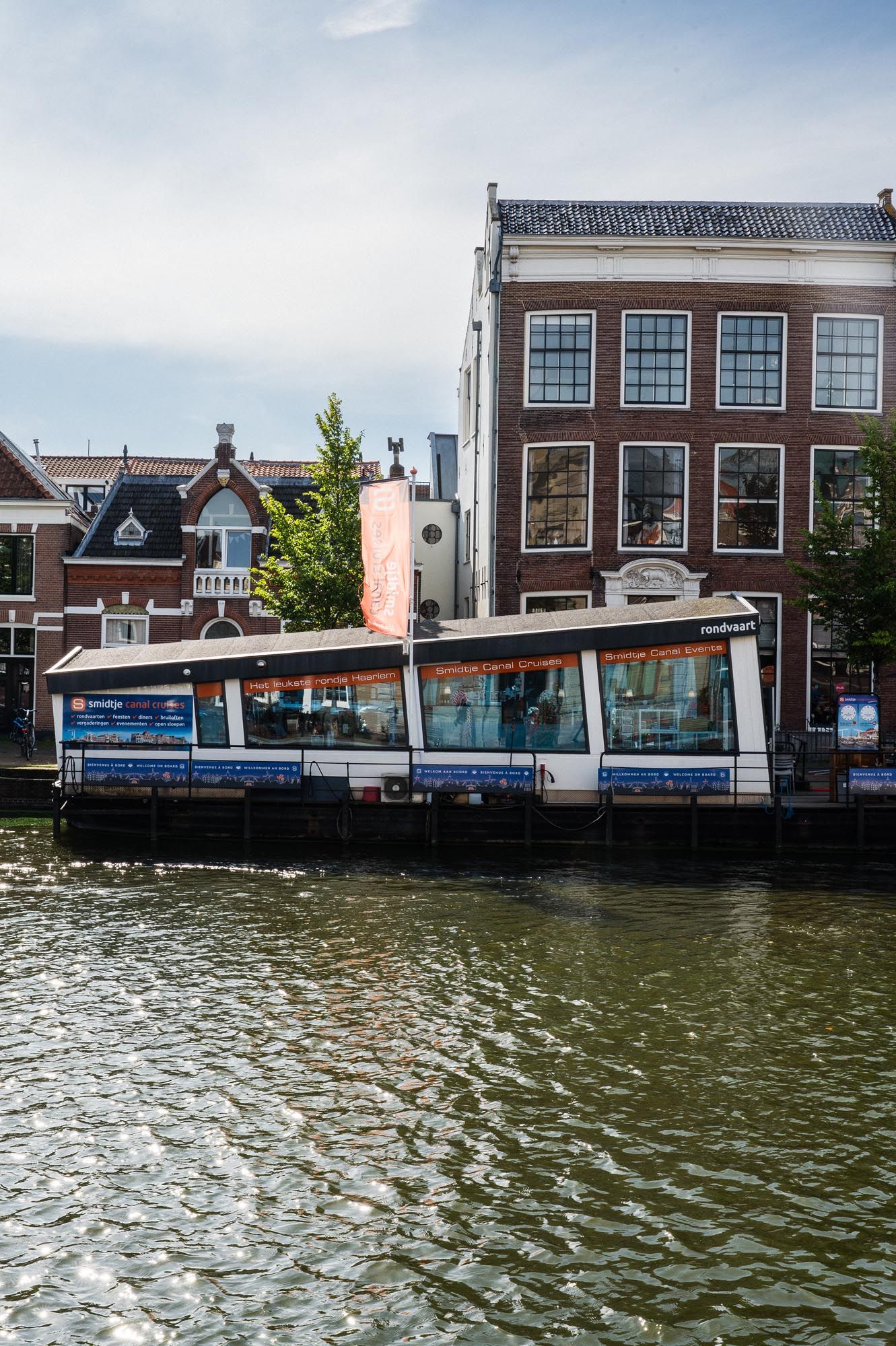 Modern ferry boat docked on Amsterdam canal with historic Dutch brick buildings in background on cloudy day.