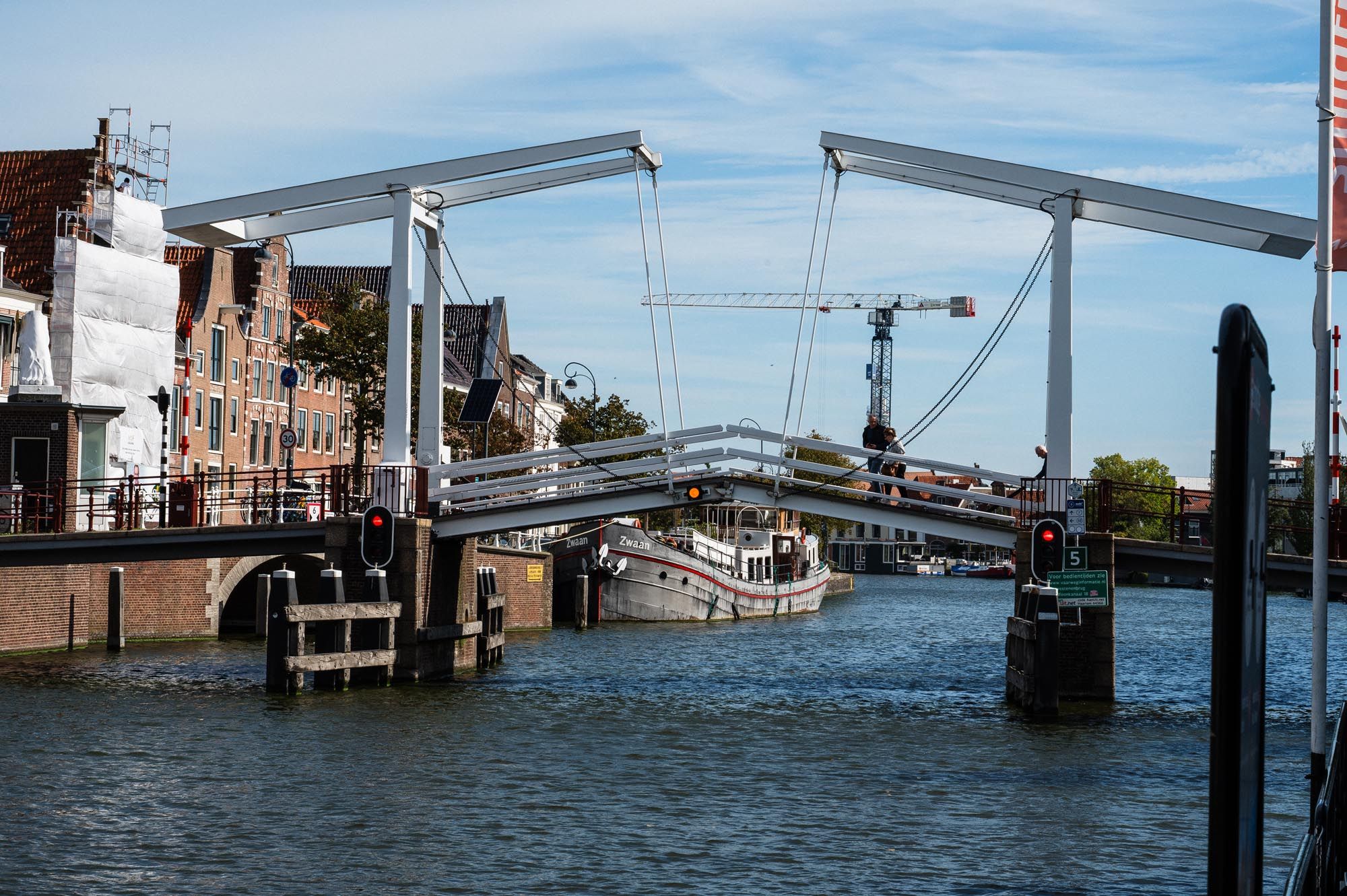 A historic Dutch drawbridge over a calm canal with boats and cranes visible under a partly cloudy sky.