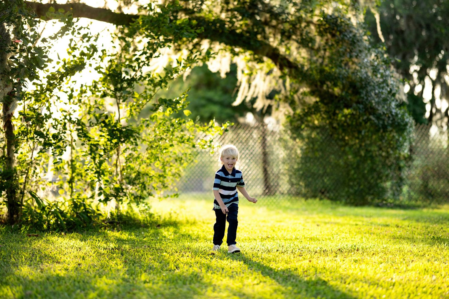 A young boy in a striped shirt runs across a lush green lawn under sunlit trees in a park.