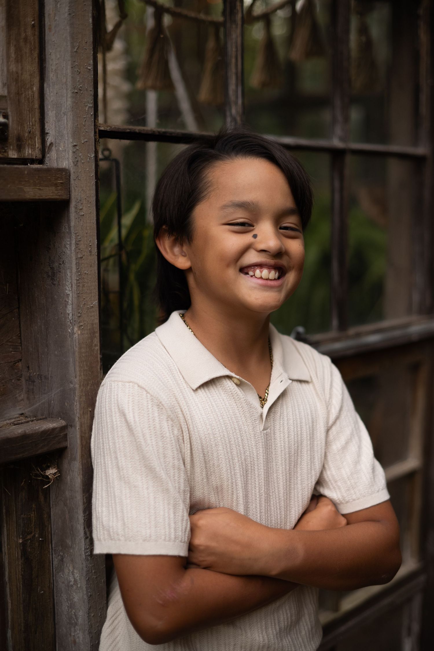 Smiling young boy with crossed arms wearing a cream polo shirt, standing against a rustic wooden door outdoors.
