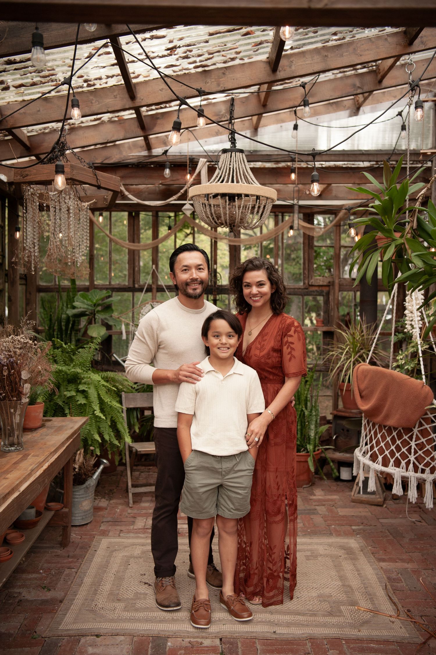 A family of three poses in a lush greenhouse with string lights and a chandelier overhead.