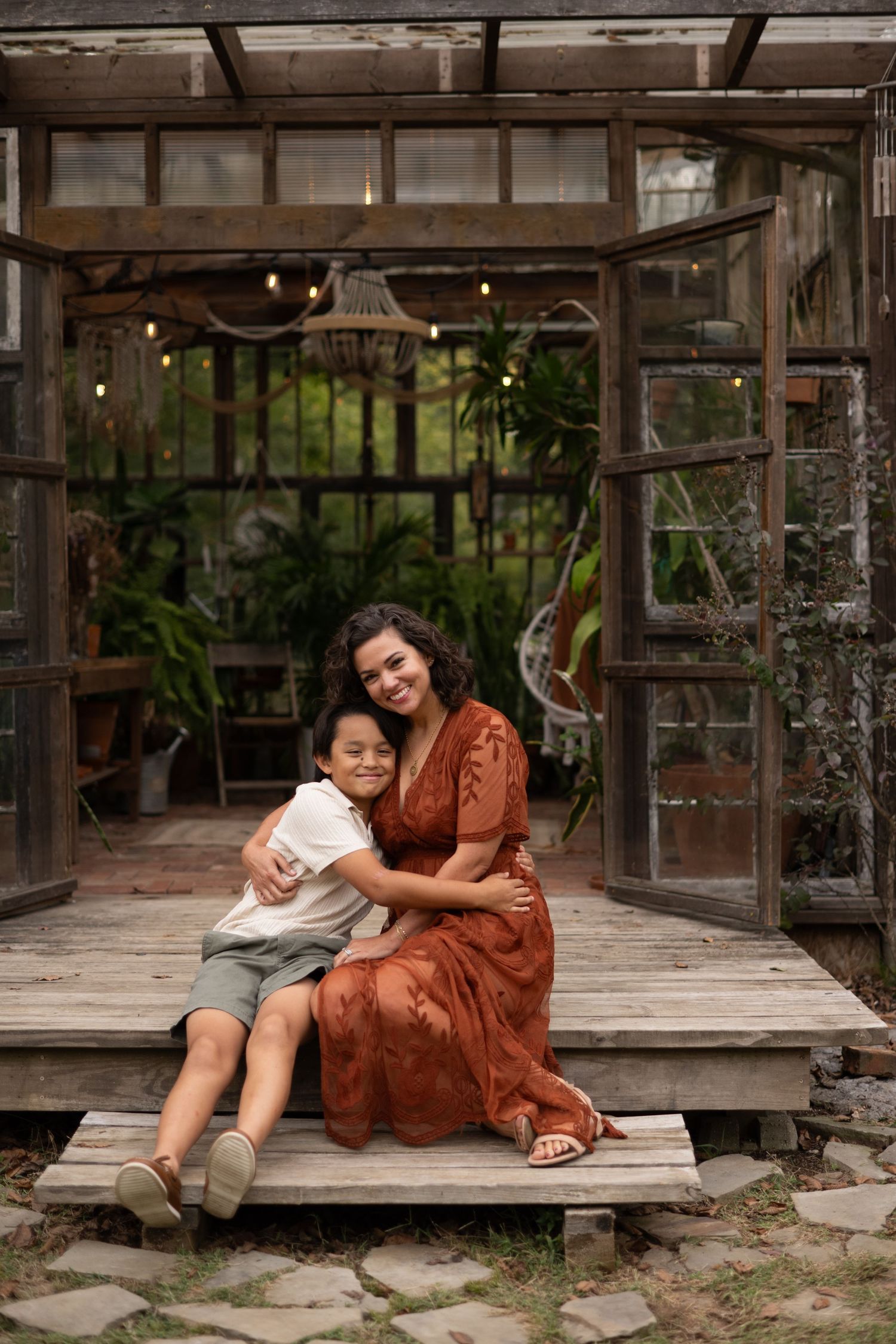 A woman in a rust floral dress hugs a young boy on a wooden porch of a rustic glass greenhouse.
