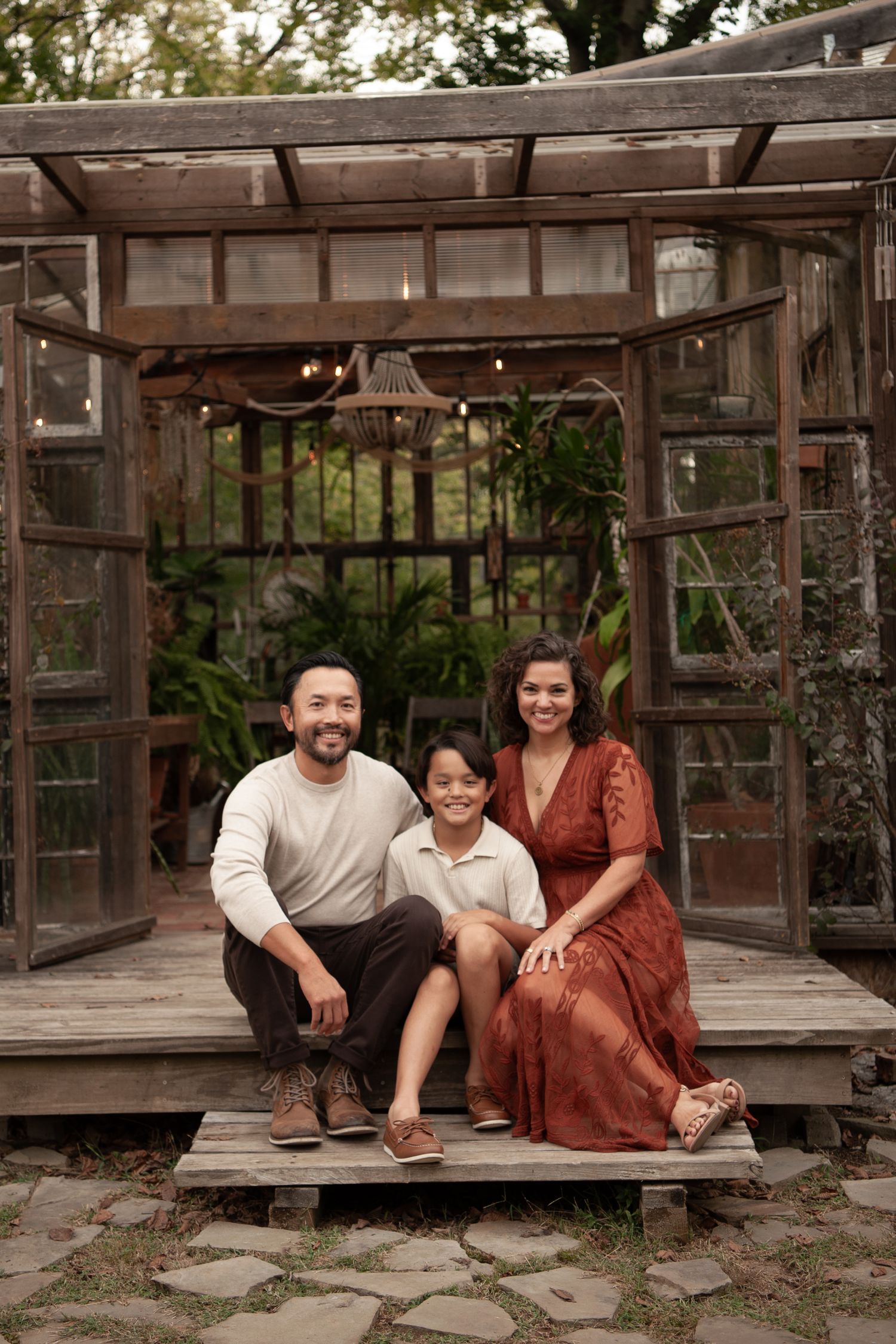 Happy Asian family of three posing together on wooden deck in front of a charming rustic greenhouse structure.