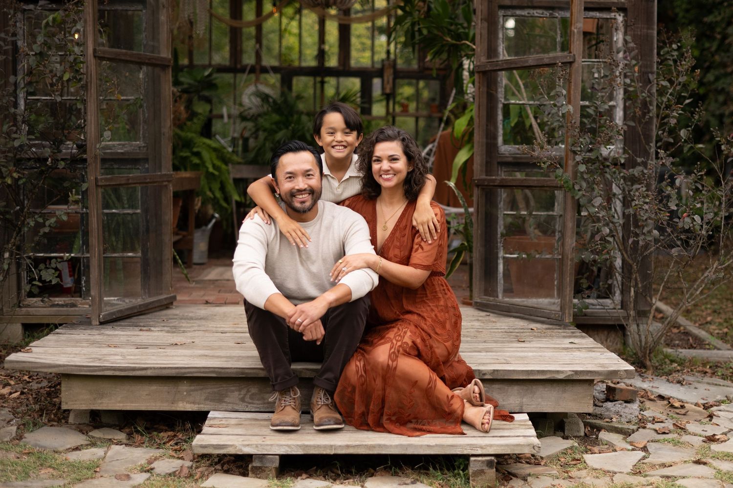 Happy family of three posing on wooden porch steps surrounded by lush garden greenery.