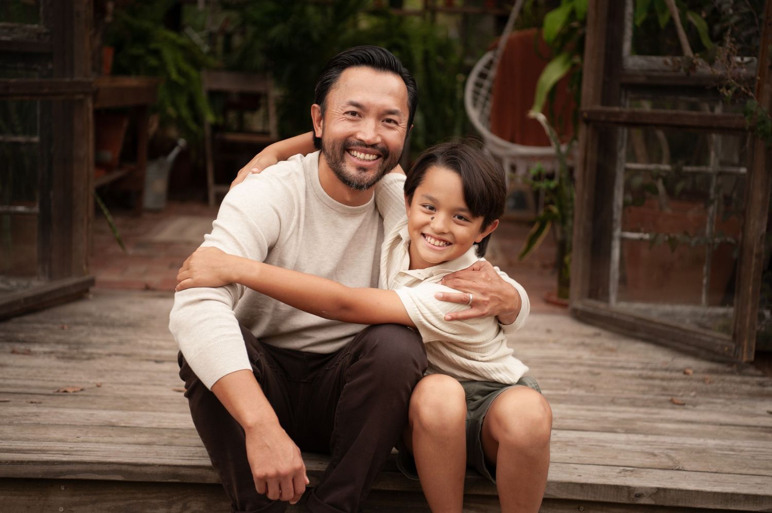 A smiling Asian man hugs a young boy on a wooden porch surrounded by lush green plants.