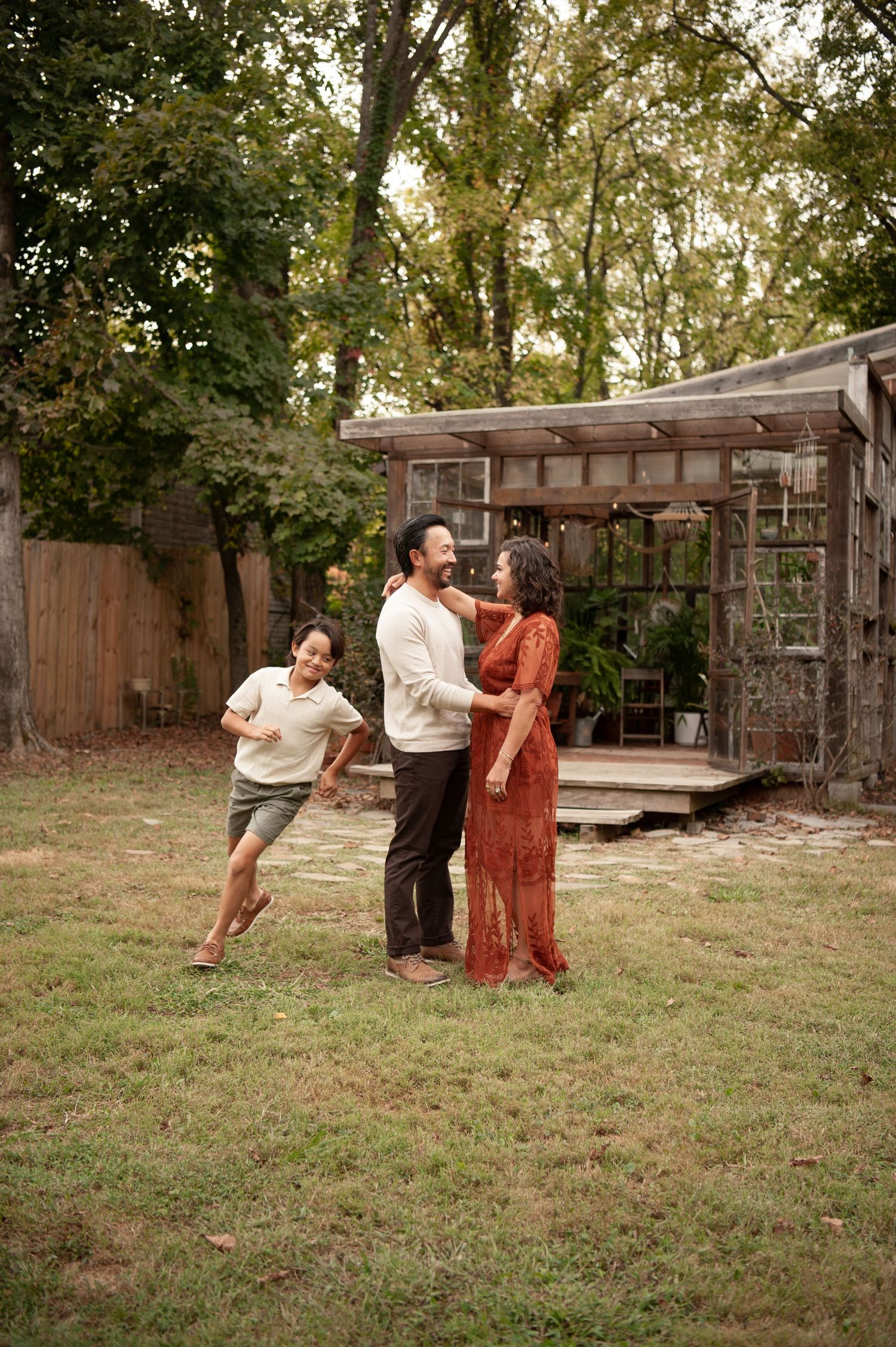 Family of three laughing and playing together in a backyard with a rustic greenhouse and tall trees in background.