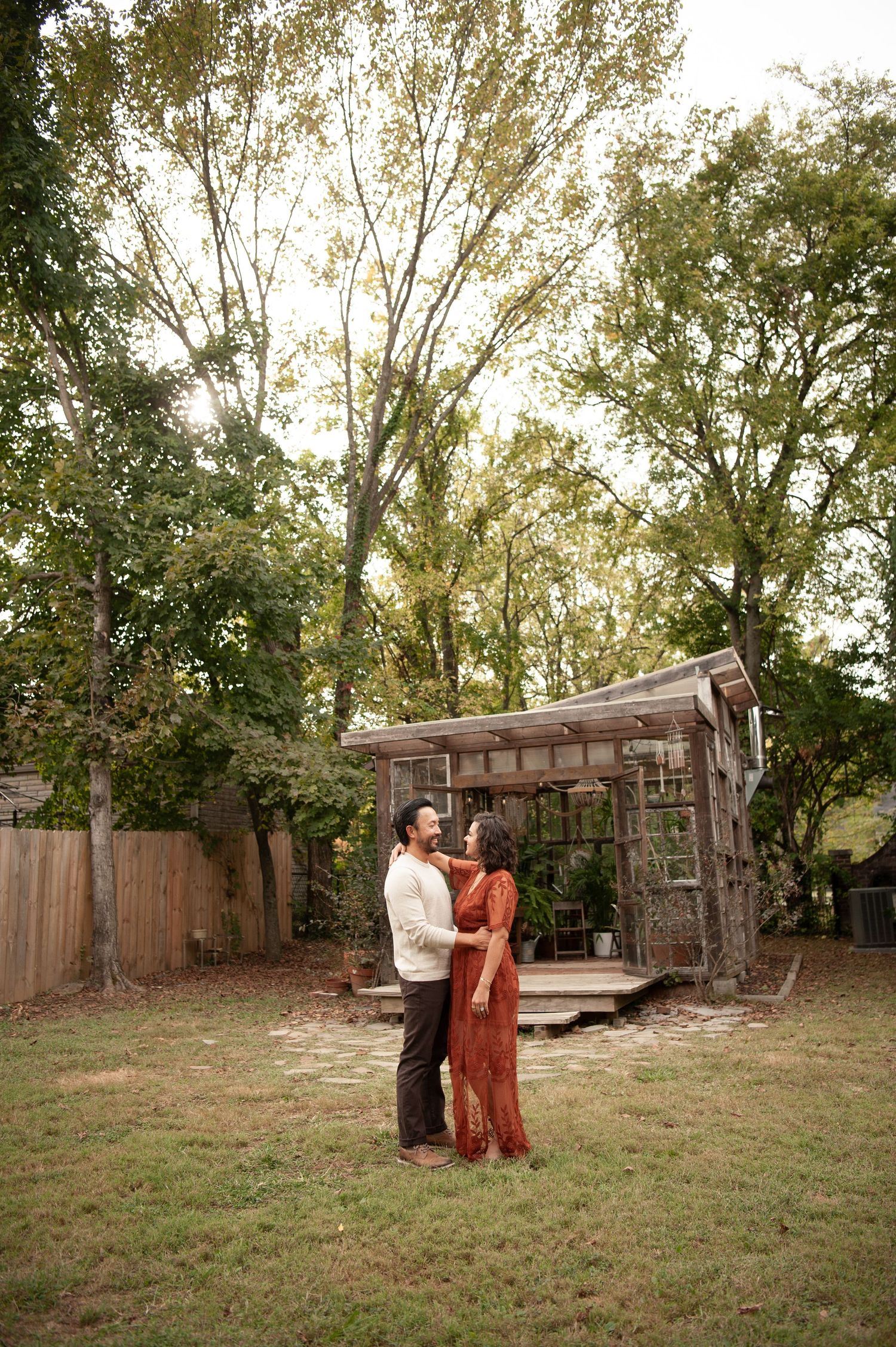 Couple in traditional attire embracing in a backyard garden with a wooden pergola and tall trees in the background.