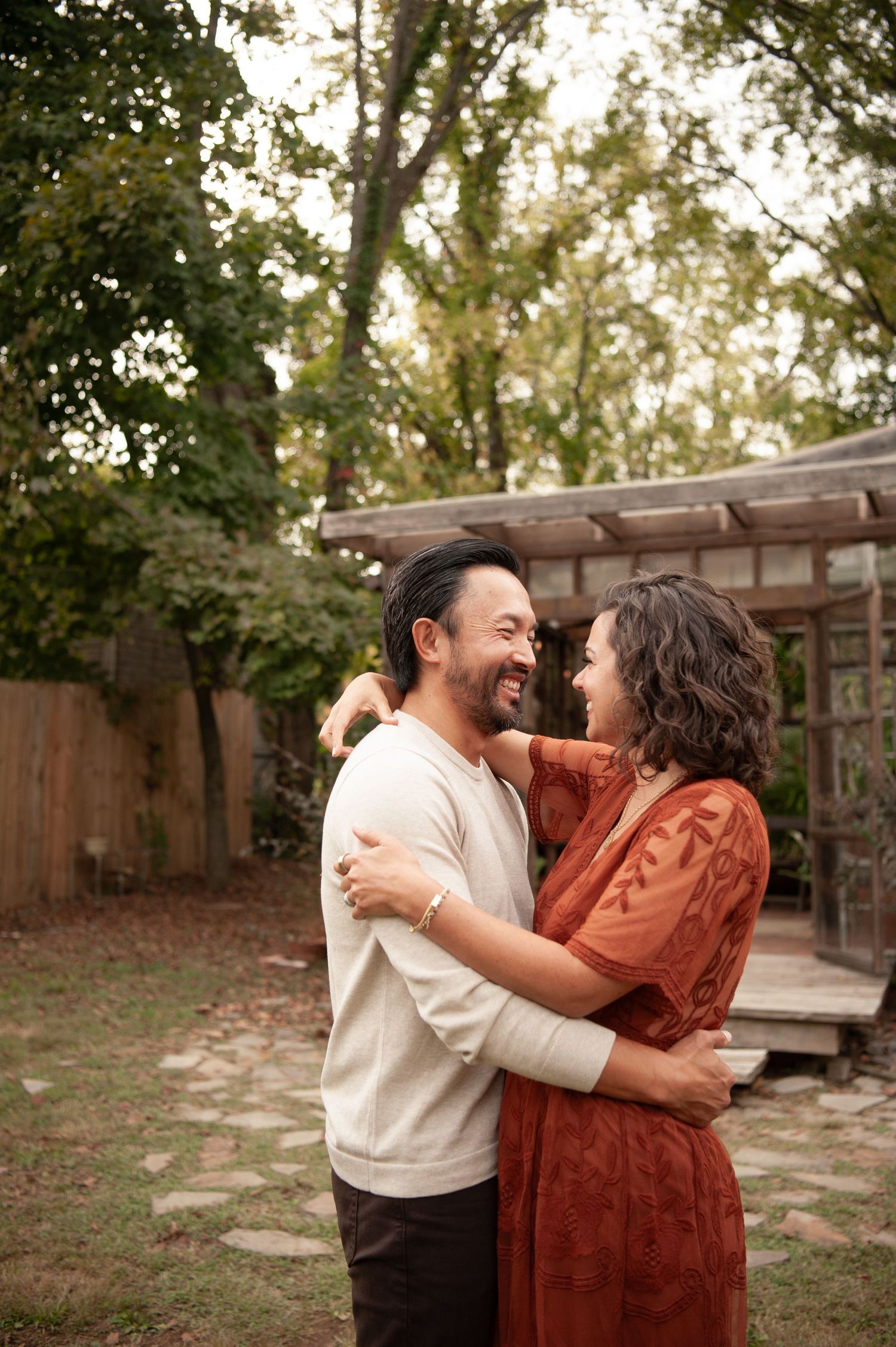 A smiling couple embraces in a lush backyard garden, the woman in a rust dress and man in a cream sweater sharing a joyful moment.