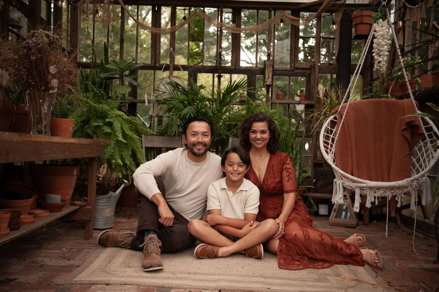 Family of three smiling together in a lush greenhouse surrounded by potted plants and hanging ferns.