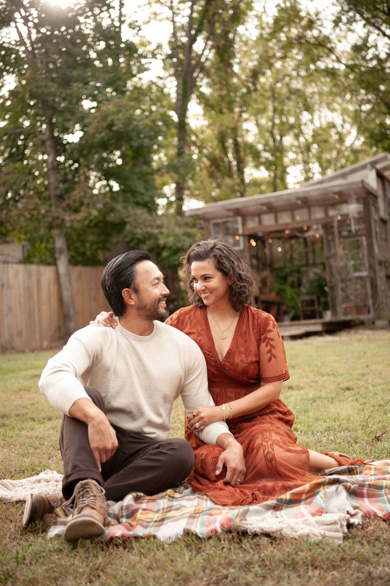 A couple sits on a plaid blanket in a backyard, smiling at each other, surrounded by lush green trees.
