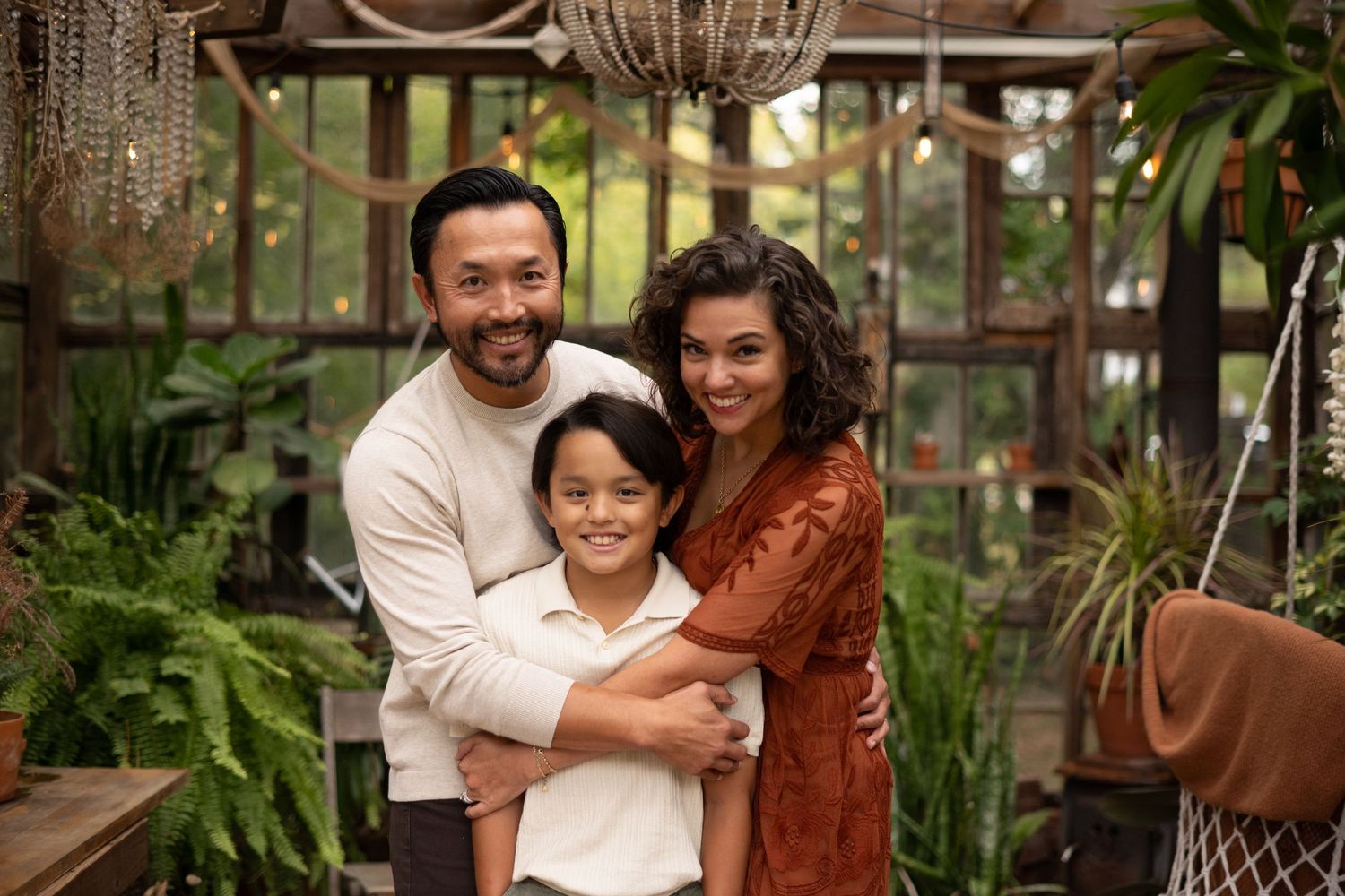A happy family of three poses together in a lush greenhouse setting, smiling warmly for a portrait photo.