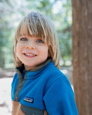 Smiling blonde child wearing a blue Patagonia fleece jacket outdoors among trees.