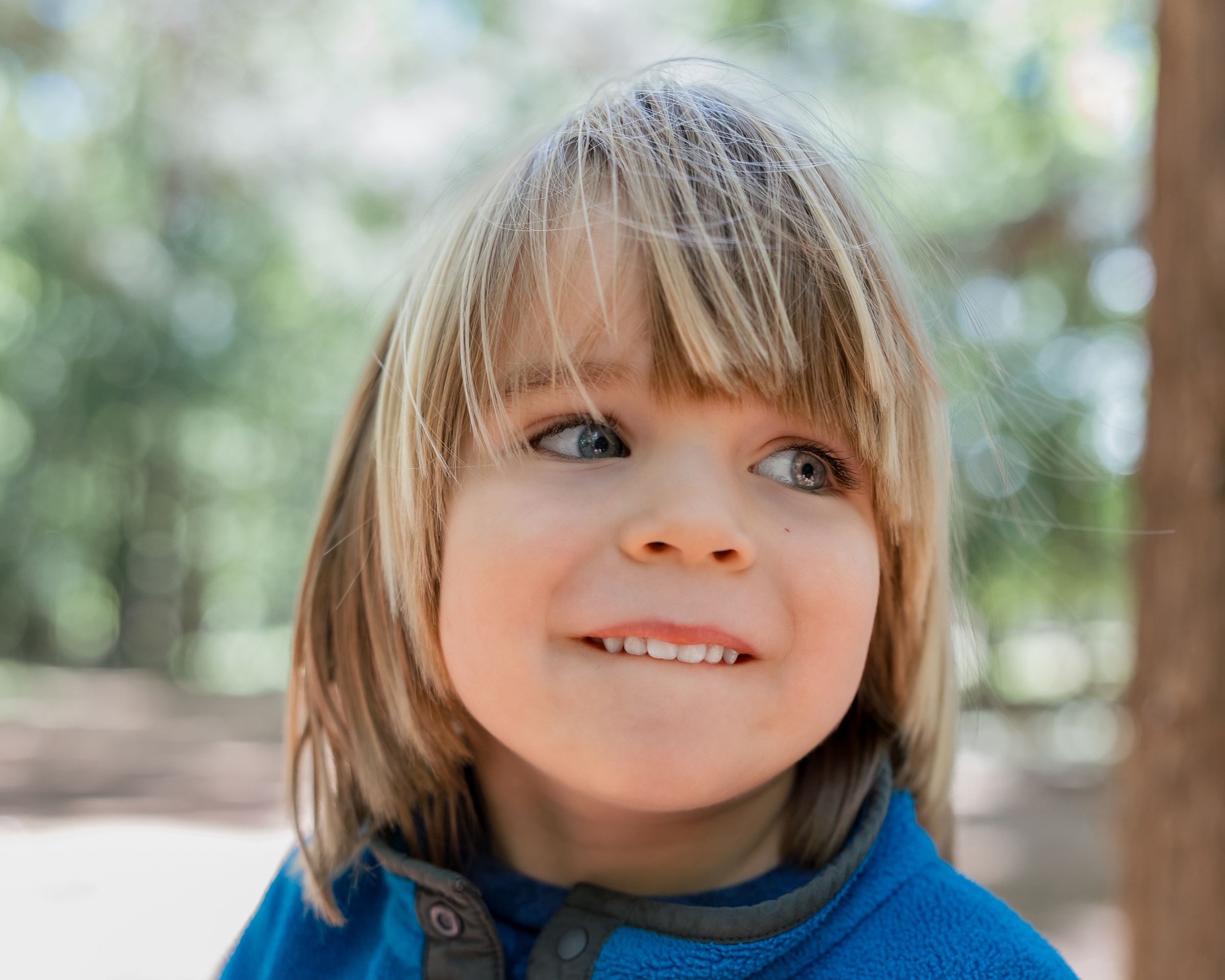 Smiling blonde child with blue eyes looking up outdoors among trees, wearing a blue jacket.