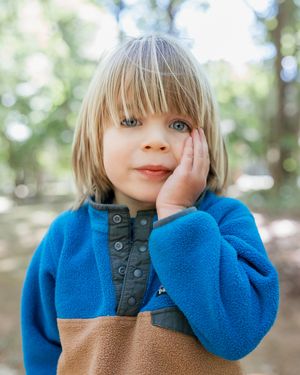 Blonde toddler in blue and tan fleece pullover rests hand on cheek, posing outdoors among trees.