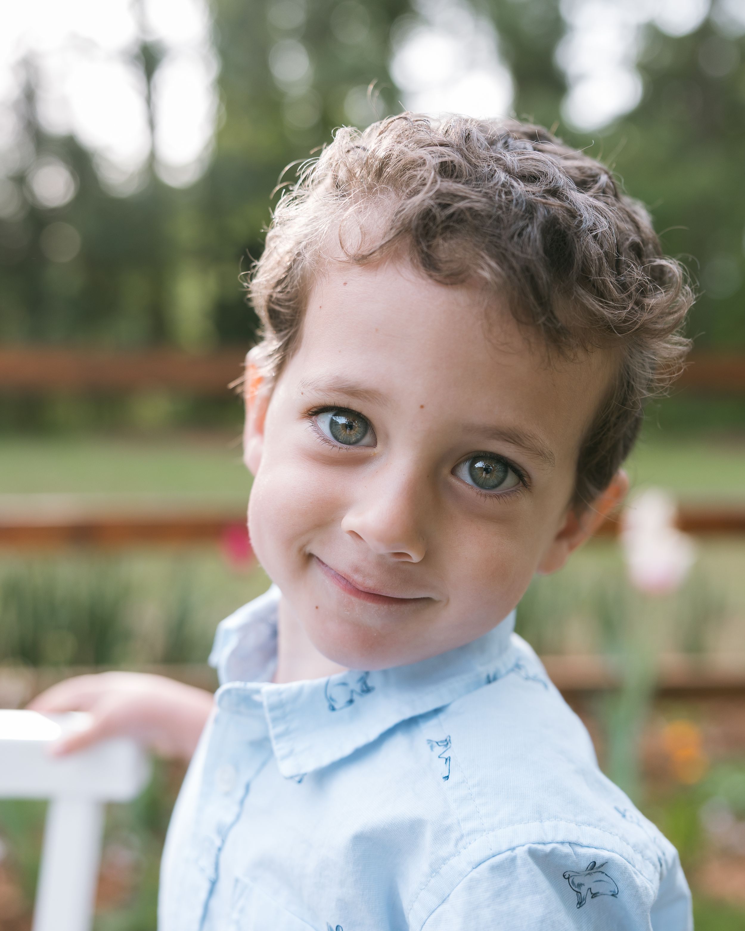 Curly-haired boy with blue eyes smiling outdoors in a light blue shirt, with a blurred garden background.