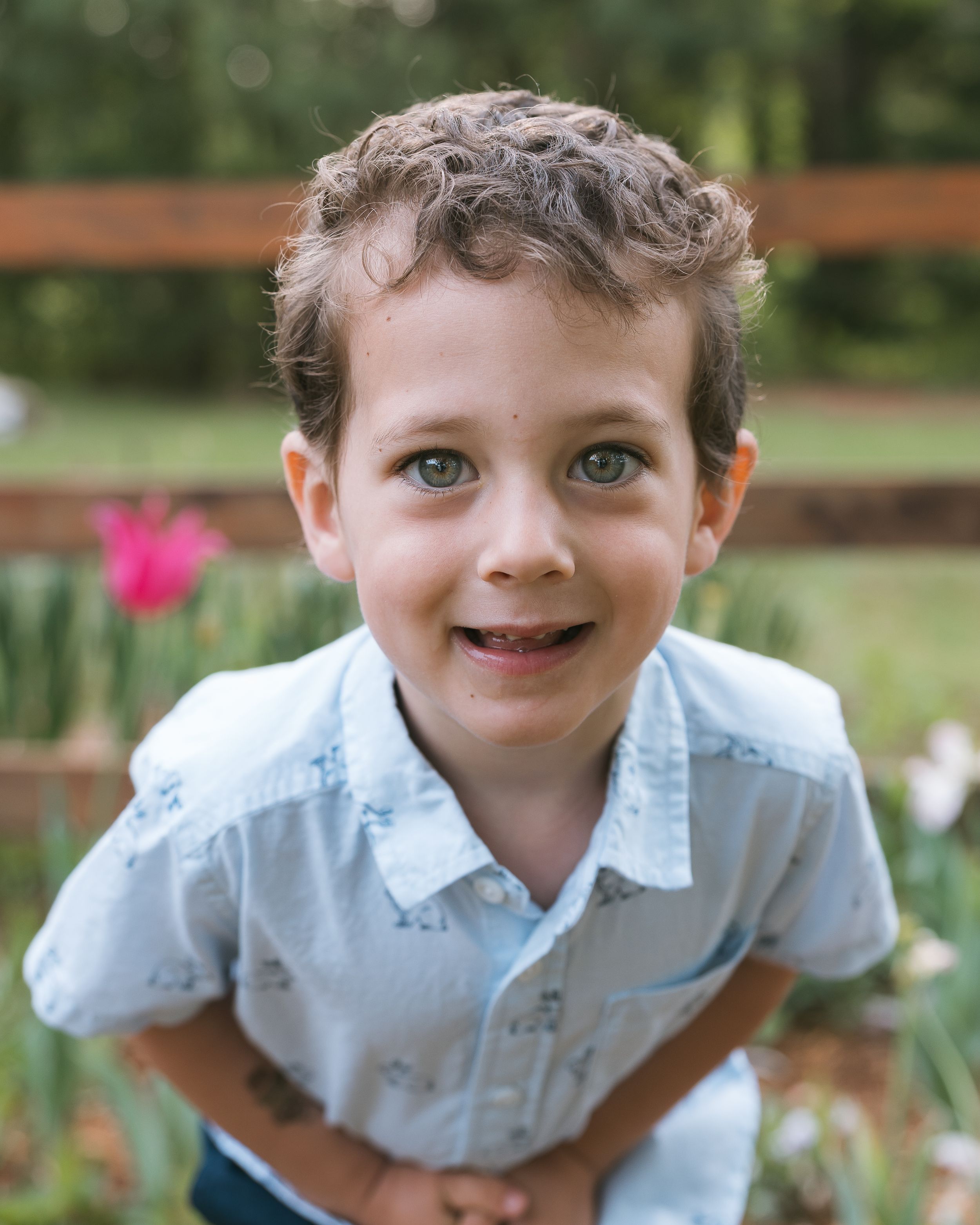 Smiling young boy with curly brown hair and green eyes wearing a light blue shirt outdoors near colorful flowers.