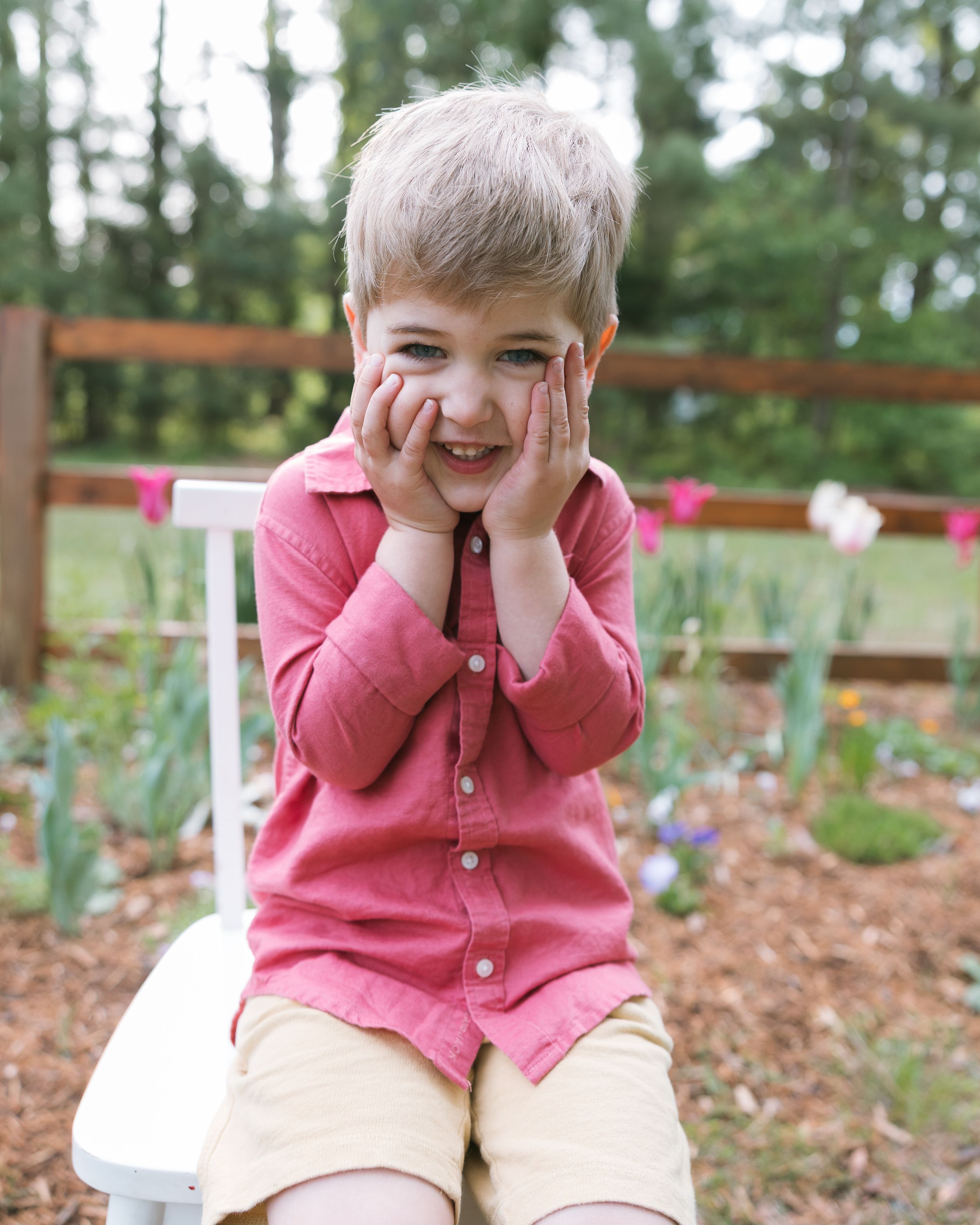 A young boy in a pink shirt sits outdoors, smiling with hands on cheeks, surrounded by a colorful garden.