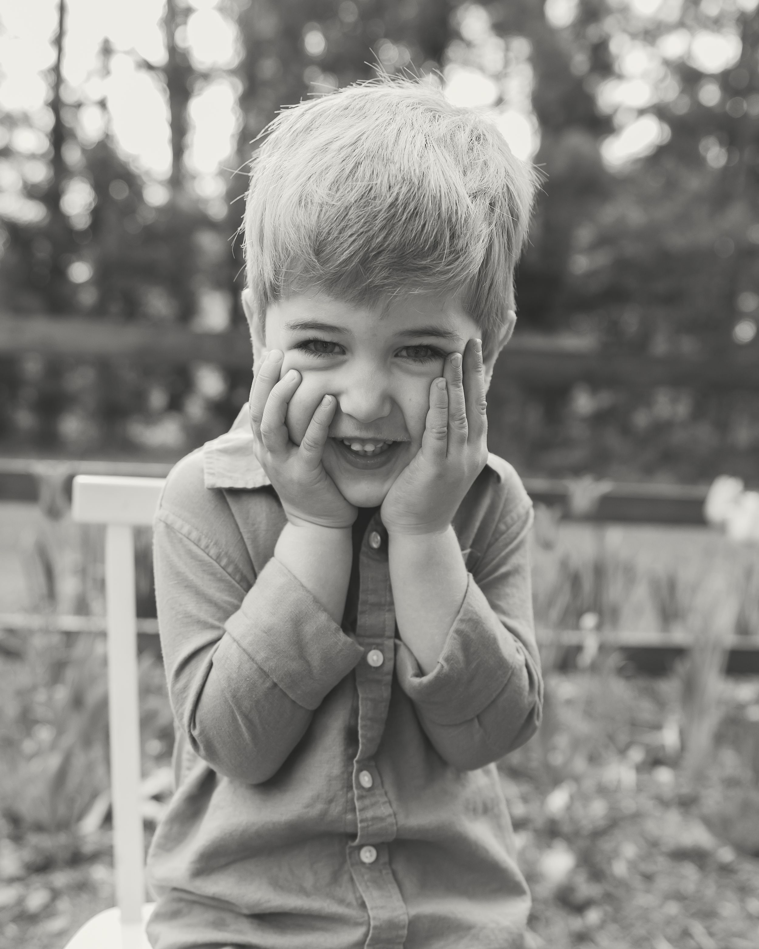 A cheerful young blonde boy squishing his cheeks with both hands, smiling outdoors in a black and white photo.