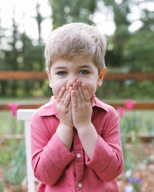 A young blonde boy in a pink shirt covers his mouth with both hands while standing outdoors in a garden.
