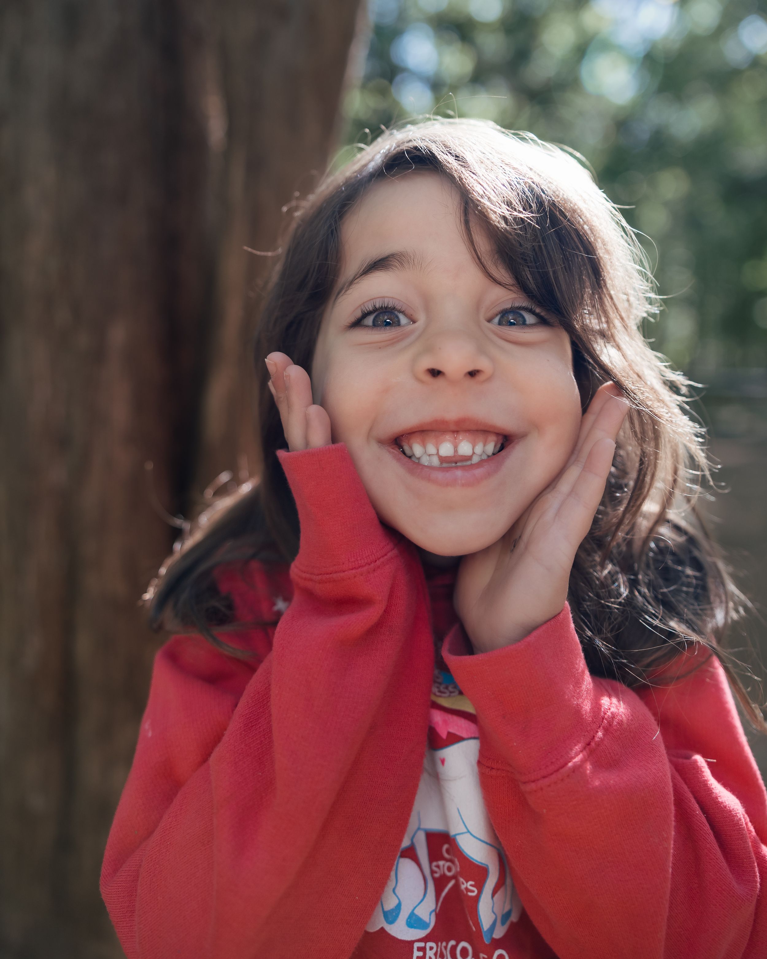 A cheerful young girl in a red hoodie smiles broadly with hands on cheeks, posing outdoors near a tree.