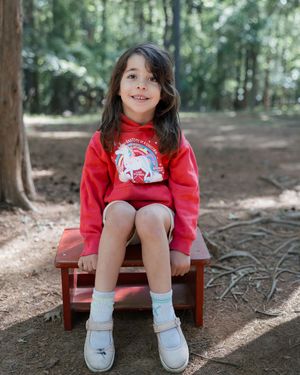 A young girl in a red unicorn sweatshirt and shorts sits smiling on a small red bench in a forest.