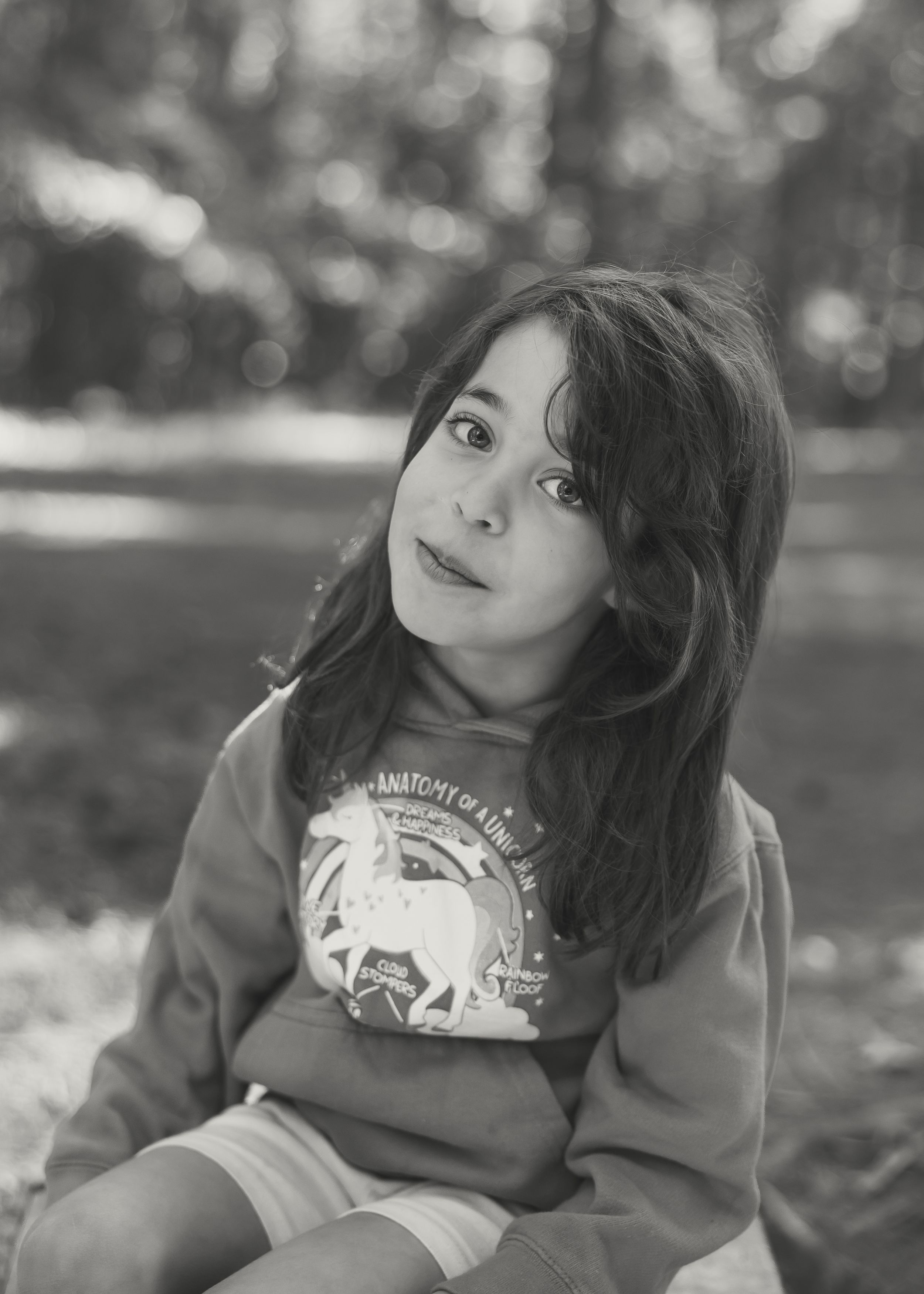 Black and white portrait of a young girl with long hair wearing a unicorn sweatshirt, sitting outdoors.
