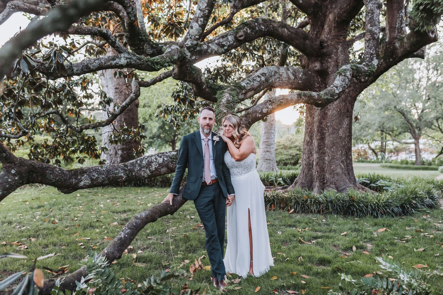 Couple embracing in a park beneath a large moss-draped oak tree, the man in a dark suit and the woman in a white dress.