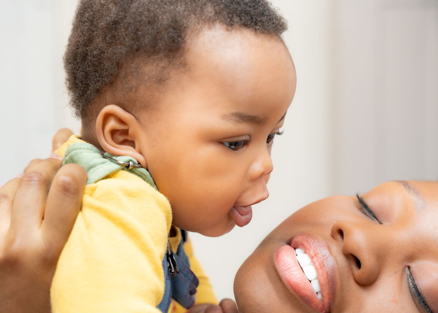 Close-up of a smiling mother holding her baby in a yellow outfit as they look at each other, with the baby leaning towards her face.