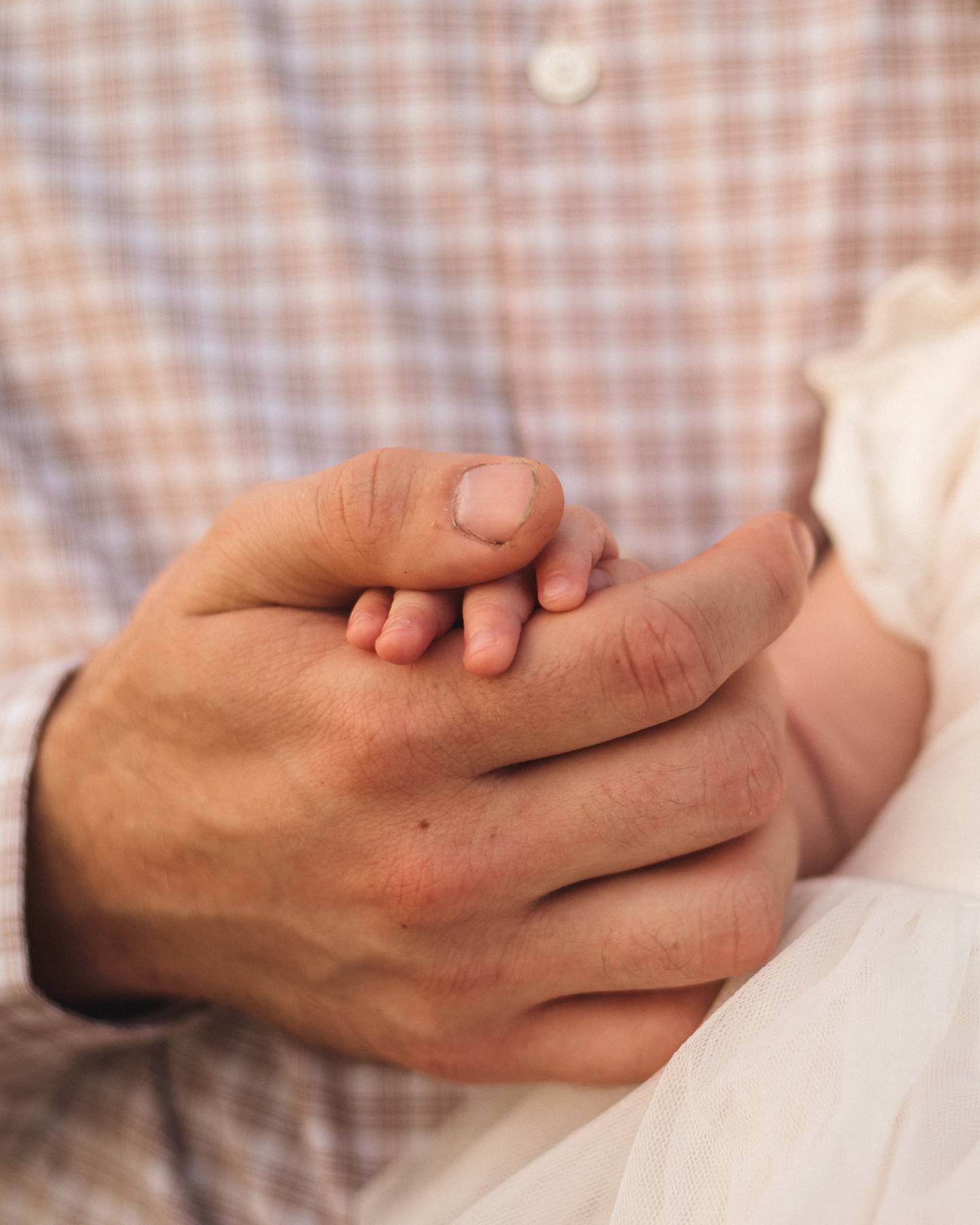 Close-up of a father's large hands gently holding a baby's tiny hand, wearing a plaid shirt in a tender family moment.