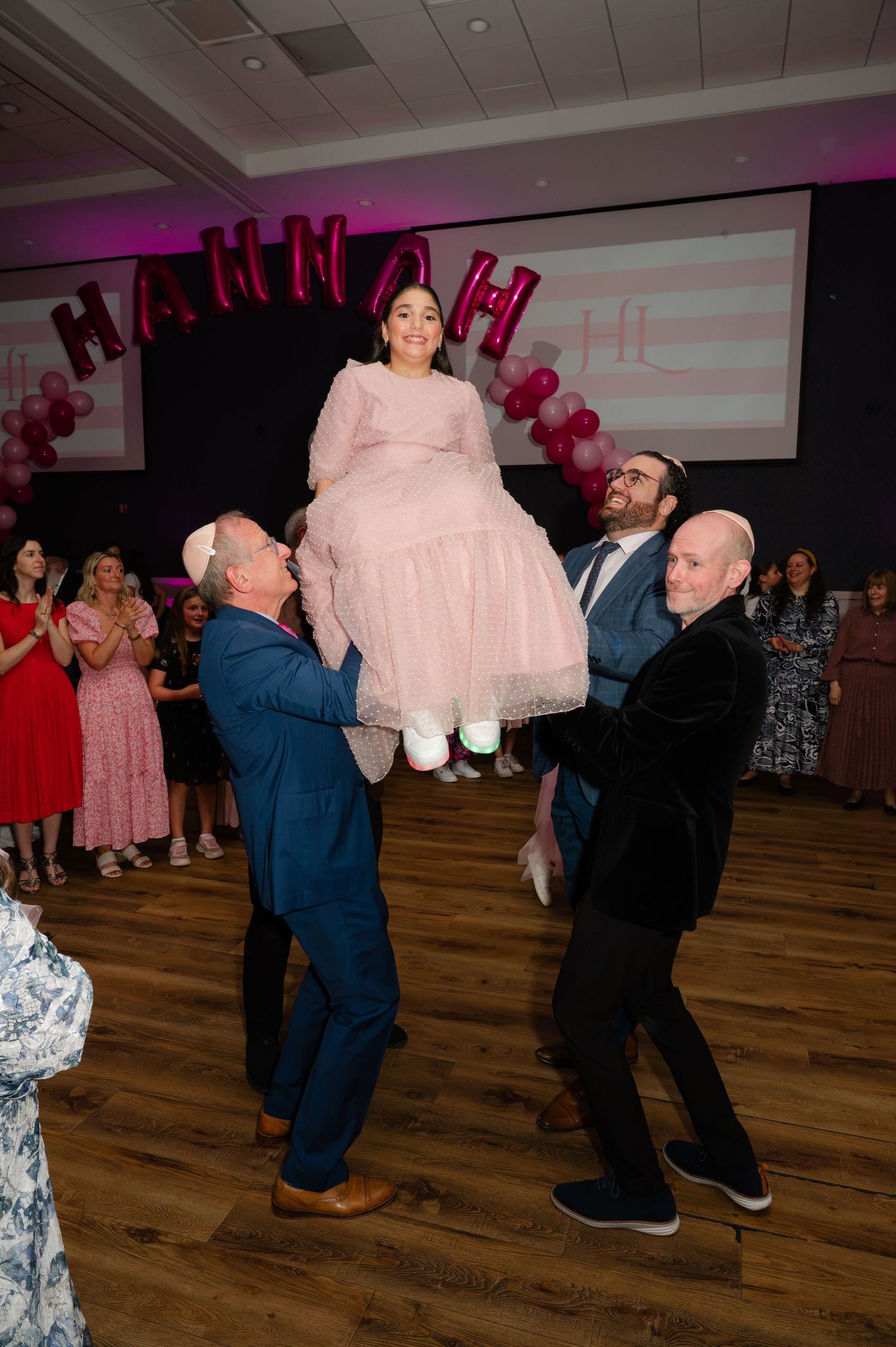 Girl in a pink dress is lifted in the air by men during a hora celebration in NYC with a balloon sign and guests cheering behind her.