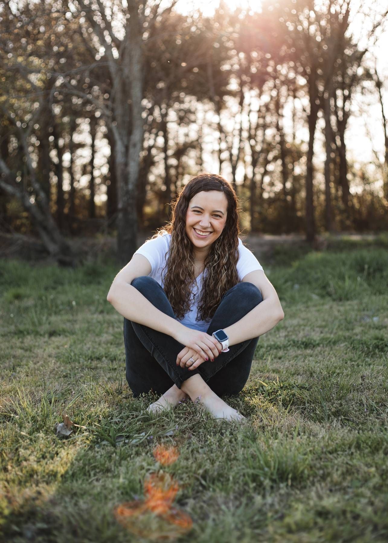 A smiling woman with long wavy hair sits cross-legged on grass in warm golden sunlight with trees behind her.