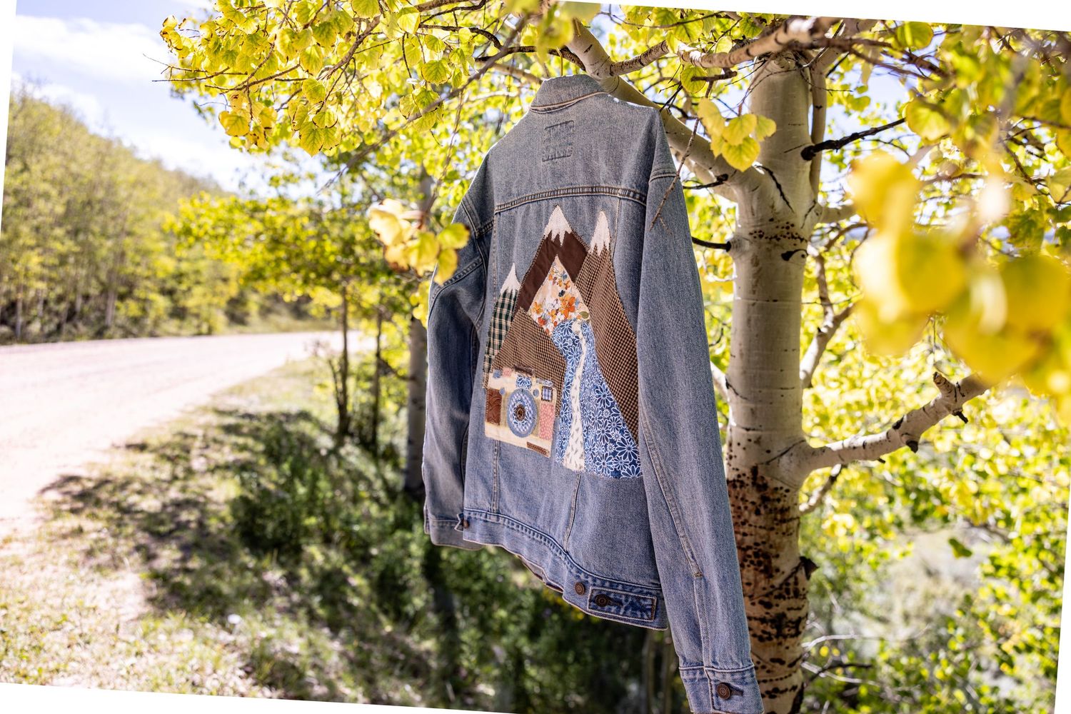 Denim jacket with a mountain patch design hanging from an aspen tree branch with golden fall leaves beside a country road.
