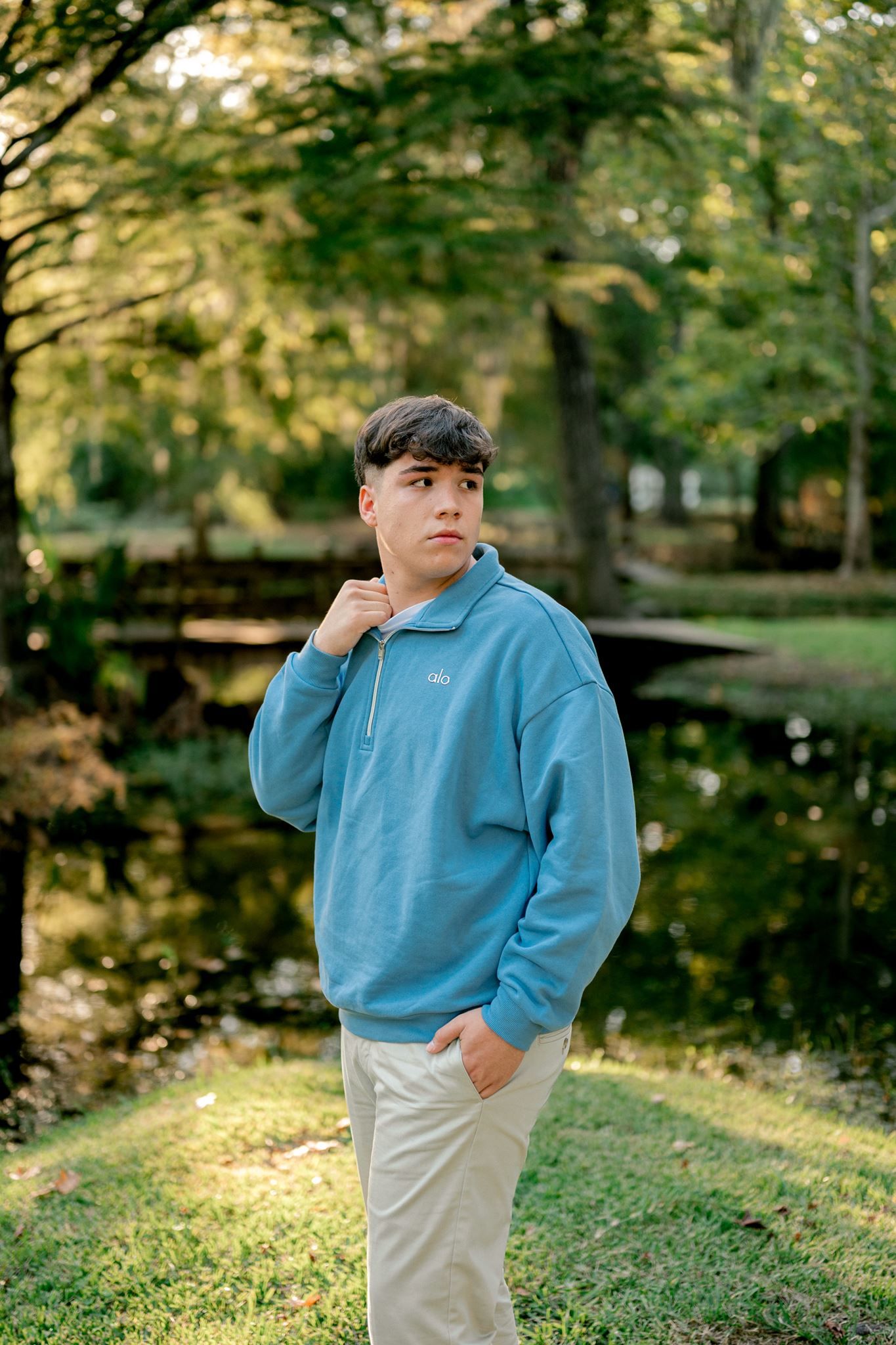 Young man in a blue quarter-zip pullover and khaki pants stands on a grassy lawn beside a pond surrounded by tall trees.