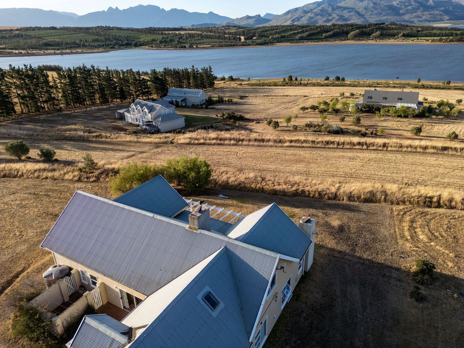 Aerial view of a gray metal-roofed farmhouse with outbuildings on dry grassland overlooking a large lake, with mountains in the distance.