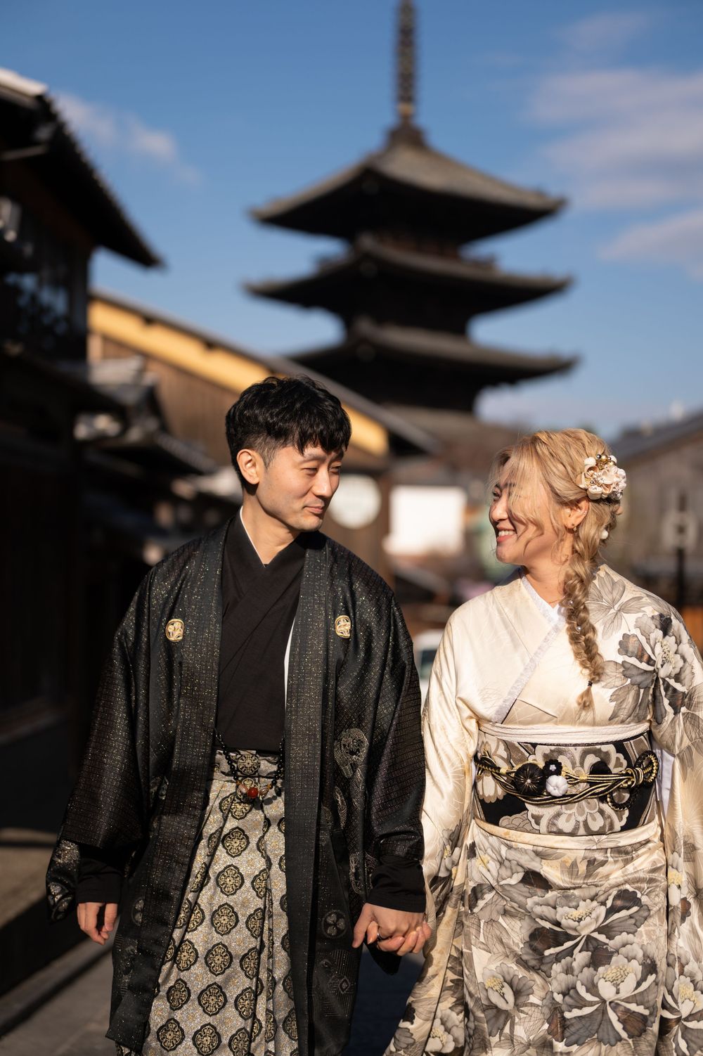 Smiling couple in traditional Japanese kimono holding hands on a Kyoto street with a historic pagoda rising behind them.