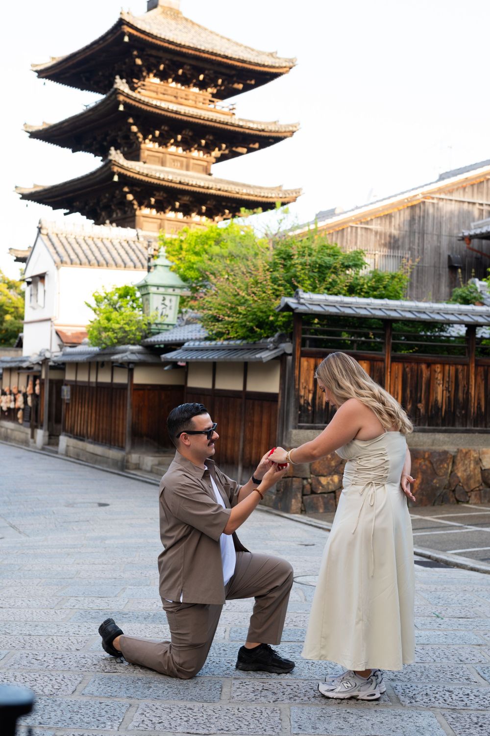 Man in sunglasses kneels on one knee proposing to a woman in a cream dress on a Kyoto street with a historic pagoda behind them.