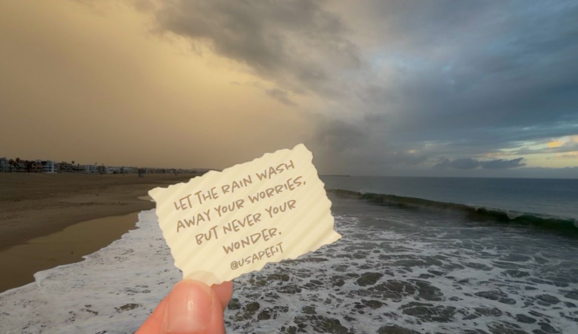 Hand holding a handwritten note reading let the rain wash away your worries but never your wonder against a stormy beach and ocean waves.