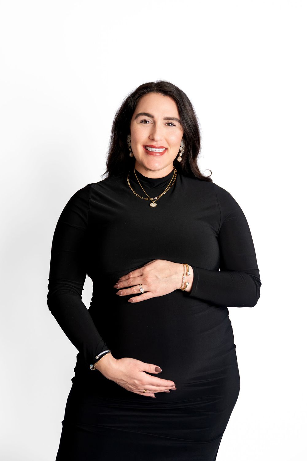 Smiling pregnant woman in black long-sleeve dress holding baby bump against white studio background.