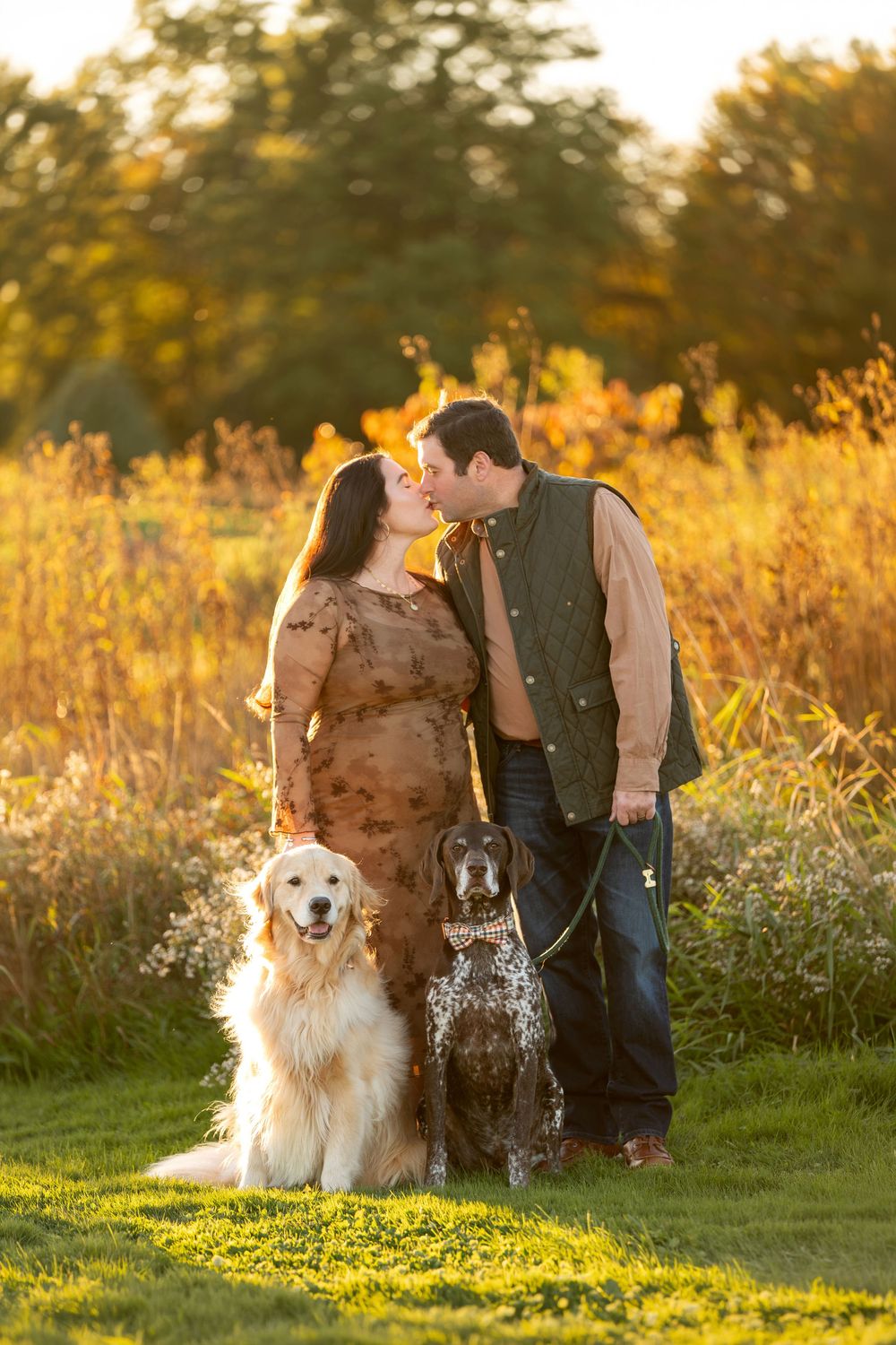 Couple kissing at sunset with golden retriever and spotted dog during outdoor maternity session.