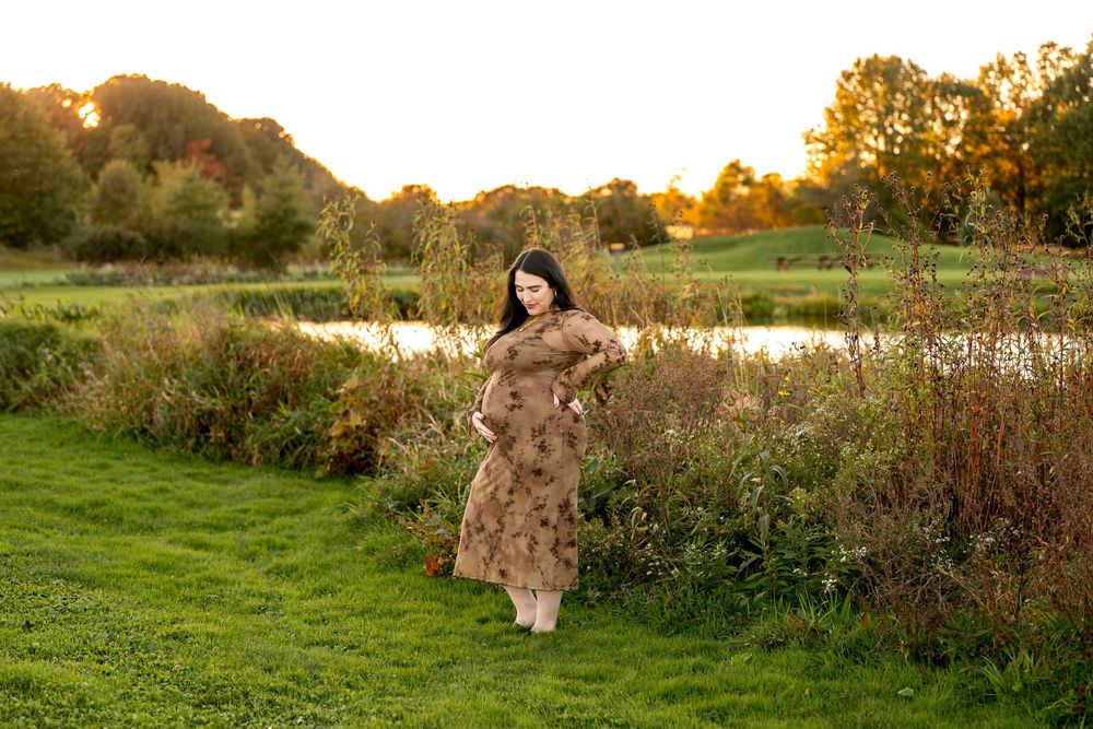 Pregnant woman in a floral dress standing by a pond in a lush green field at golden hour.