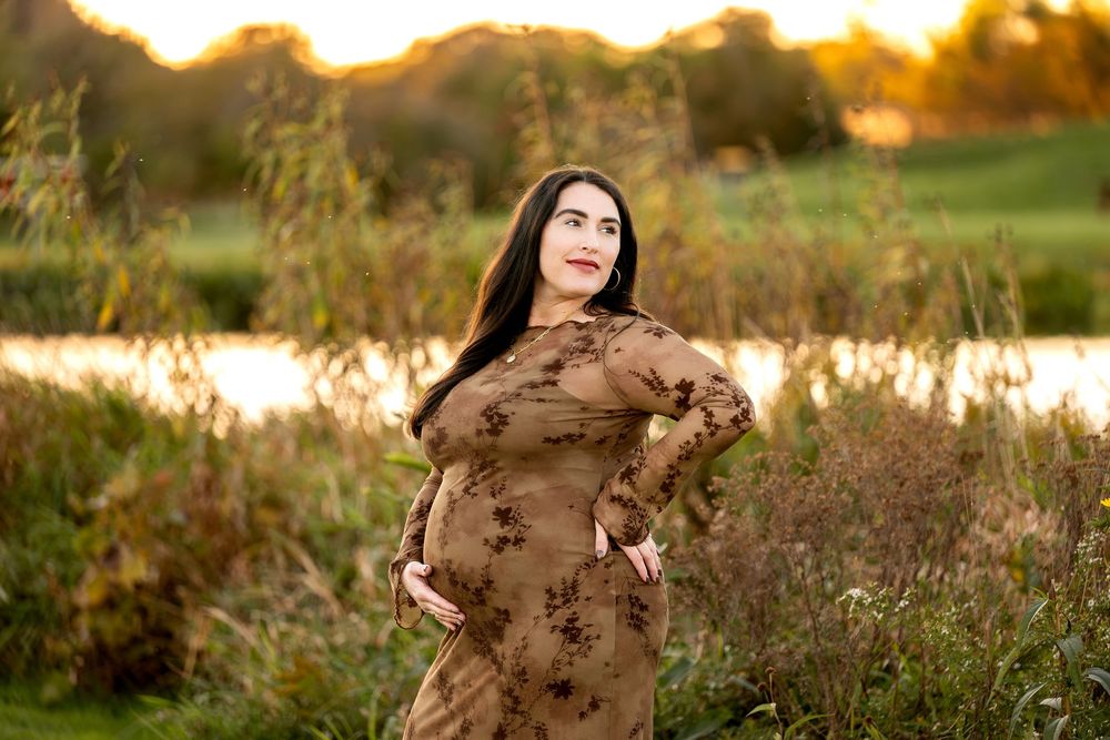 Expectant mother in brown floral dress posing outdoors by a pond during sunset.