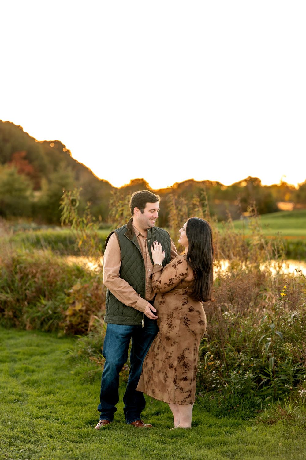 Couple sharing a tender moment by a pond at golden hour during fall maternity photography session.
