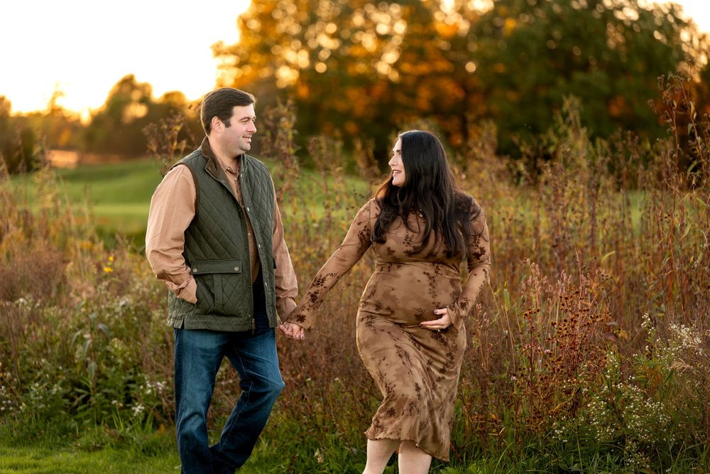 Couple holding hands in autumn field at golden hour, pregnant woman in brown dress cradling baby bump.
