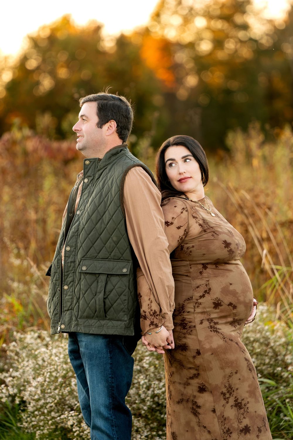 Couple holding hands back-to-back in autumn field, pregnant woman in brown floral dress at golden hour.