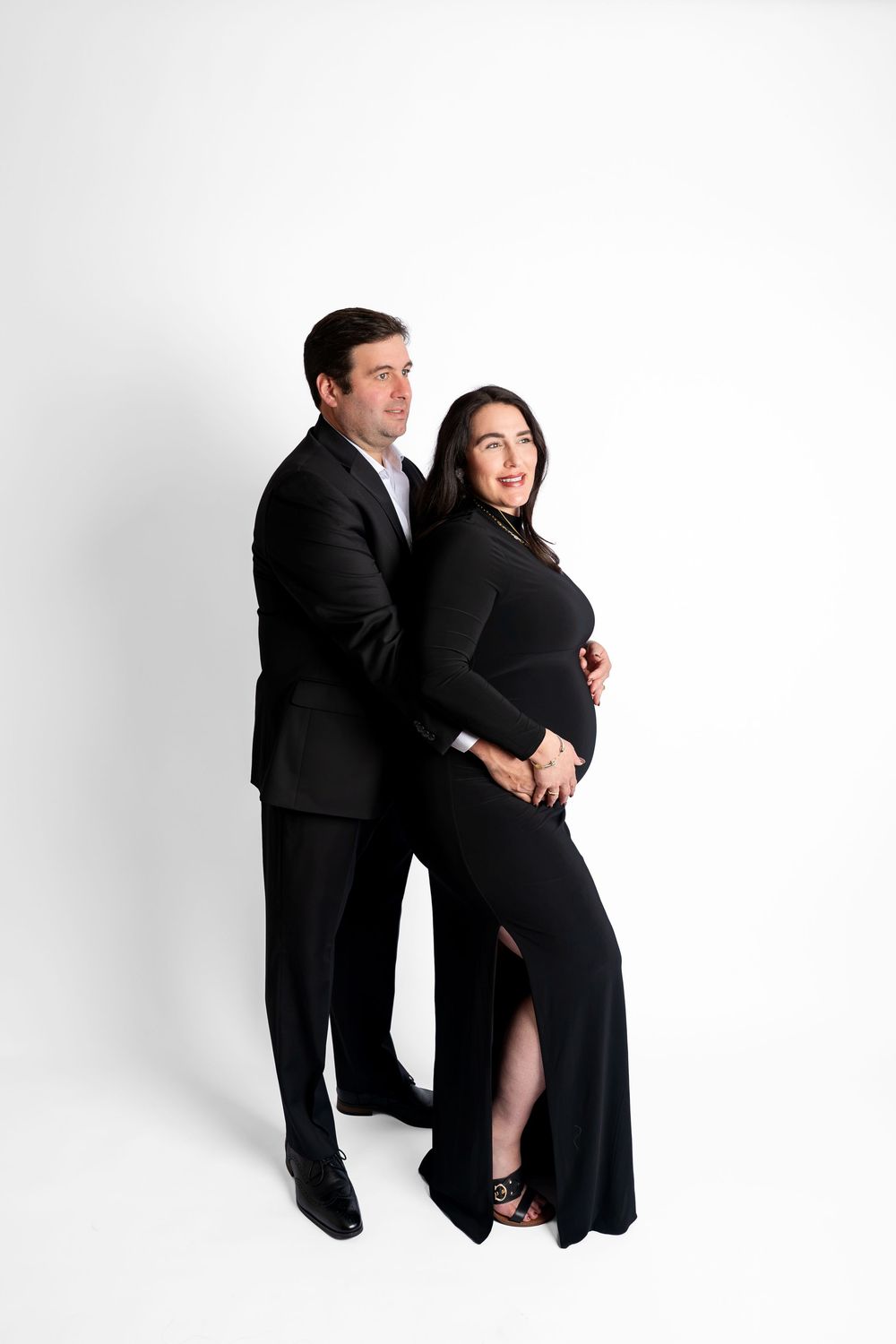 Elegant maternity photo of a couple in black formal wear against a white studio background.