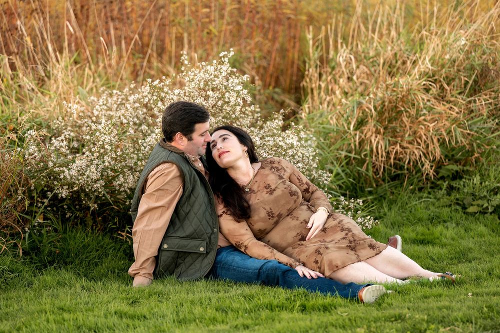 A couple sits on grass, the woman leaning back on the man as he kisses her cheek, surrounded by wildflowers.