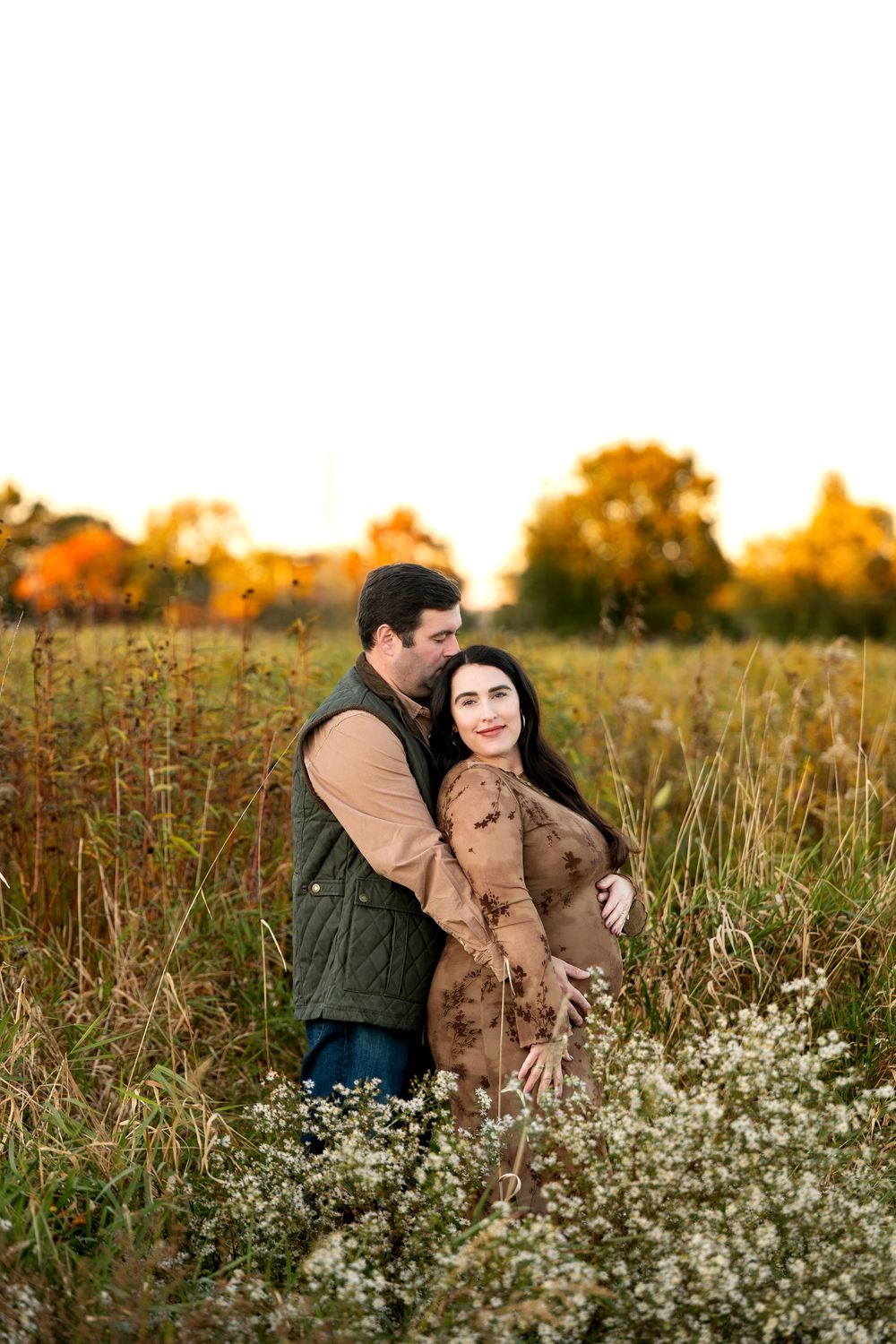 Pregnant couple embracing in autumn field with golden wildflowers during sunset maternity shoot.